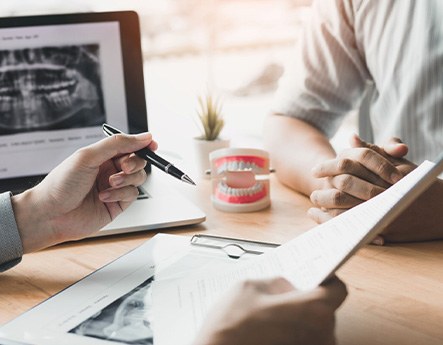 Hands of dentist and patient at desk with X-rays, model teeth, and treatment plan